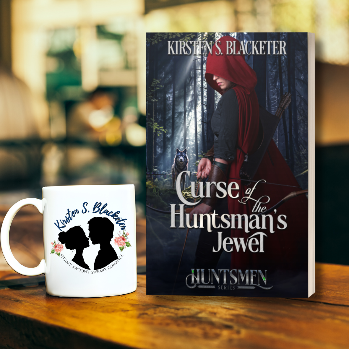 Paperback book "Curse of the Huntsman's Jewel" with dark forest cover and red-cloaked woman holding a bow with a wolf in the background. Beside the book is a white coffee mug with the author's logo on it featuring a silhouette of a couple.